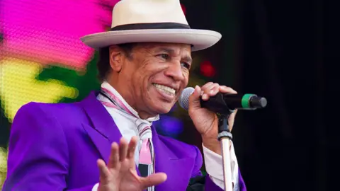 Redferns/Getty Images Kid Creole performing in 2015. He is wearing a purple suit with a pink tie and white hat. He is holding onto the microphone while singing into it.
