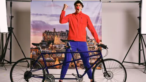 Greg James standing behind a tandem bicycle wearing blue trousers, a red top baseball cap and a red nose on his nose.