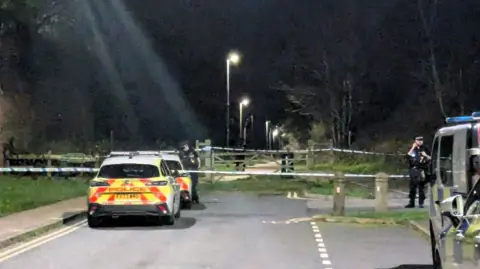 BBC Three police cars and a police van parked on either side of an empty road. Police officers and a cordon can be seen in the distance