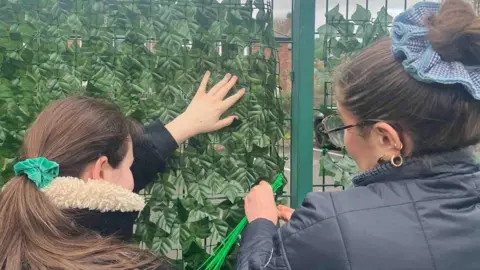 Fauve Walkerdamarell Two young women stand holding zip ties and shrubbery next to a green metal fence. 