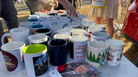 Colección de tazas de Phillip Norton/BBC dispuestas sobre una mesa. En primer plano, se observa una pila de fotos del presentador Peter Levy. Se aprecia la mano de una persona que intenta alcanzar una de las tazas.