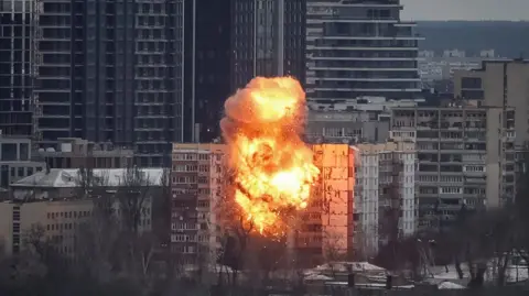 Reuters Orange fire and smoke rise from a multi-storey building after it is hit by a missile in Kyiv on 27 December. Taller buildings are situated behind the one targeted, with trees in front of it.