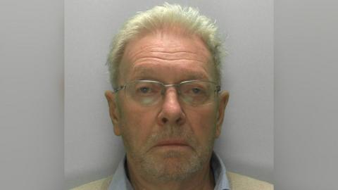A police custody image of Dr Charles Ragan, taken against a grey background. He is pictured unsmiling, wearing thin-framed glasses, a pale collared shirt and a light coloured jumper. 