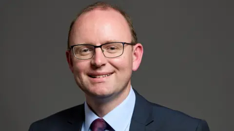 UK Parliament Andrew Cooper, the MP for Mid Cheshire, has receding brown hair and wears glasses. He smiles for the camera while wearing a dark suit, pale blue shirt and purple tie.