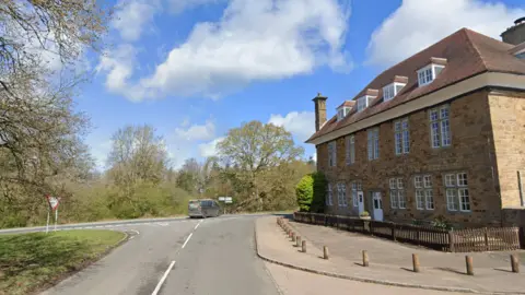 Google A 17th Century hunting lodge hotel stands at a T-junction in the Forest of Dean on a sunny day. A van can be seen turning right. The junction is surrounded by trees.