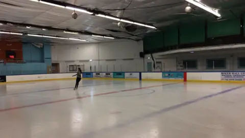An ice rink with barriers with adverts on them around the side. You can see one female ice skater turning around the rink.