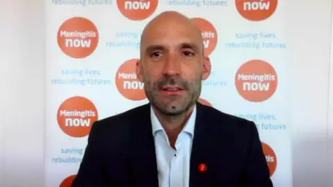 A man with a shaved head and short facial stubble appears on a video call in front of a branded Meningitis Now backdrop. He is wearing a dark suit jacket over a white collared shirt, with an orange charity pin attached to the lapel.