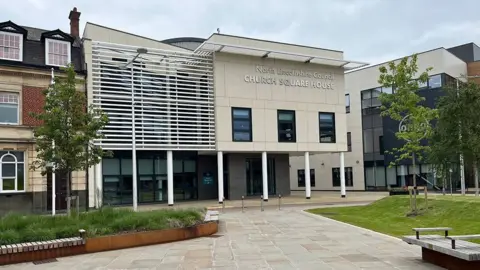 LDRS Exterior of North Lincolnshire Council's offices in Scunthorpe a two-storey building clad in light stone with a lattice on one side and windows on the ground floor facing out onto a paved square with plants and wooden seating.