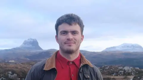 Matthew is wearing a red shirt and brown jacket with a wide collar. Behind him are mountains and sky. 