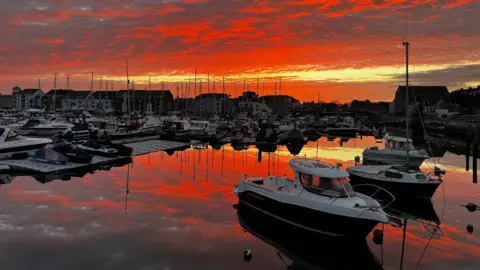 Jarnie A red and orange cloudy sky is reflected in the water of a boatyard.