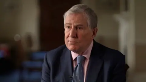 A head-and-shoulders portrait of Bruce Houlder, a white man with grey hair, wearing a navy suit, a pink shirt and a blue tie, looking to the side of the camera with the background out of focus