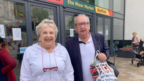 Naj Modak/BBC Jan and Alan Birch have white hair. They have lilac tops and he has a blue blazer. They both have glasses although hers are on a chain around her neck. They are outside a glass building.