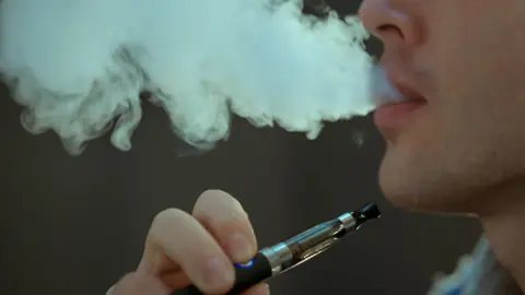 Getty Images A close-in picture of a young man smoking a vape and blowing out a cloud of light grey smoke