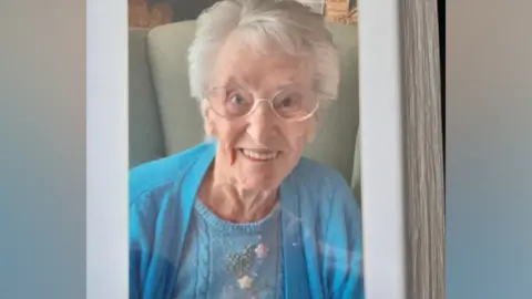 Handout Joyce Moulton pictured sitting on a chair smiling. She has grey hair and is wearing a blue cardigan and glasses.