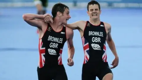 EPA Two men with short brown hair run on a blue background. They are wearing black triathlon suits with white writing that reads: Brownlee GBR