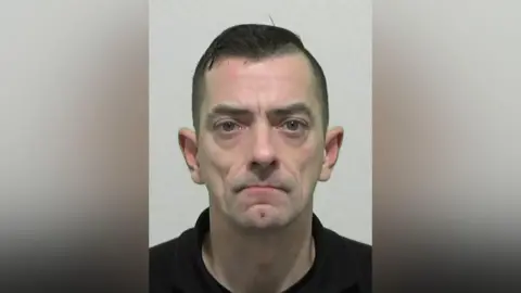 Mugshot of Mark Williamson. He has a tall thin face, with short black hair and is wearing a black polo shirt.