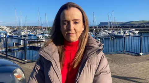 Cara McShane is standing near a pier and is wearing a purple coat and red jumper. 