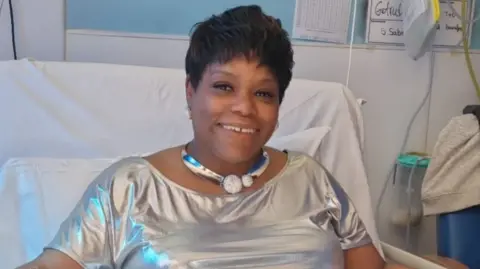 Althia Bryden in a hospital bed, wearing a silver top and large necklace and smiling at the camera