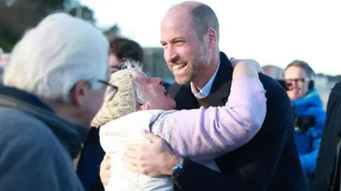 PA Media A older woman with white hair, wearing a pink jumper, gilet and a woolly hat, embraces Prince William as she looks up at him. Prince William smiles as people around them watch.