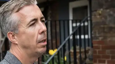 A man in a grey top looks away from the camera in mid-conversation with someone out of shot. He has grey hair and is standing in front of a home with steps leading up to its front door