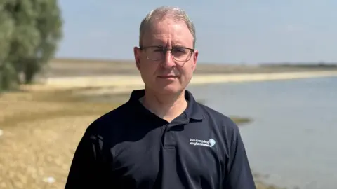 Janine Machin/BBC Geoff looks at the camera with a fairly serious expression. He wears a navy blue collared casual shirt with the Anglian water logo that says "Love every drop".