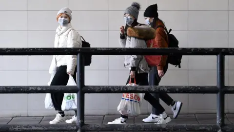 Three women walking through Newcastle city centre. They are walking on a pavement behind a railing. They are wearing hats, thick coats and facemasks.