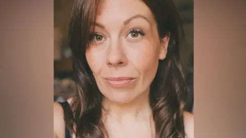 A woman with dark brown hair looks directly at the camera with green/brown eyes, a smile and a small nose ring.