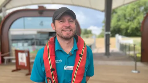 Martin Giles/BBC A head and torso shot of Tobi Radcliffe. He has a short trimmed light brown beard and is wearing a red buoyancy aid over a turquoise polo shirt as well as a black cap. He is smiling and standing outside.