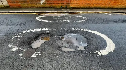 The image shows a pothole in the middle of a road, with some dirty water in them.