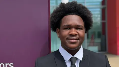 A head and shoulders picture of DJ Mudziviri who is wearing a black suit jacket and tie over a white shirt. He is standing in front of a college. He is looking and smiling directly into the camera