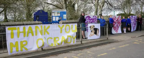 PA Media Pupils and teachers from Aldington Primary School pay their respects to Paul O'Grady with picture collages of their drawing of dogs along the route of his funeral in the village of Aldington, Kent ahead of his funeral