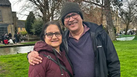 Meena Kumari Meena Kumari's parents, smiling at the camera. Her mum is wearing glasses and a purple jacket, with her dad wearing a dark jacket, purple jumper. There are trees in the background and green grass.