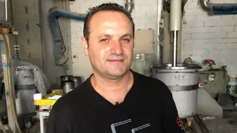 BBC Dimitris Koumatsioulis is pictured in a factory setting, with industrial drums visible in the background