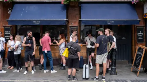 A group of people stand on the pavement drinking outside The Blue Posts pub in London. The pub has blue awnings over the windows with The Blue Posts written in gold above them