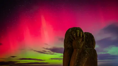 The northern lights have created a striking line of pink across the sky. Starlight is poking though. There is a moss-covered grey statue in the foreground.
