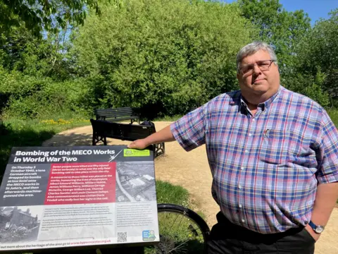 Richard Udall pictured in a blue and red checked shirt. He has grey hair and is wearing square framed glasses. He is leaning on a sign that reads "bombing of the MECO works in World War Two"