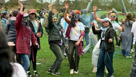 Amith Nair A crowd of people dancing during the Holi event in the park. They have many colours of powder on them. It is overcast.
