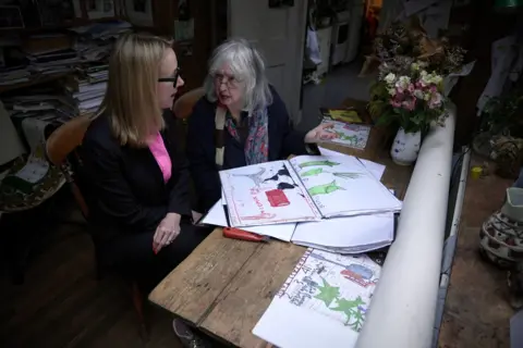 BBC/Adam Walker Katie Razzall, seated closest to the reader, in a dark jacket and pink shirt talking to Rose Wylie in a dark top and striped scarf. On the wooden table next to them are Rose's drawings - two in green, another with red, black and grey.