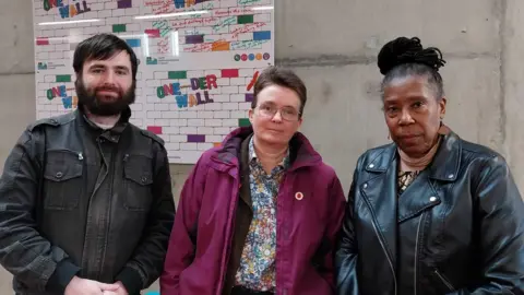 Local Democracy Reporting Service Chris Ashton, Marion Turner-Hawes and Sylvia Erskine stand side-by-side against a wall. Ashton has a dark hair and a dark beard and is wearing a black jacket. Turner-Hawes is in a purple coat and flowery shirt. Erskine is in a leather jacket and has her black hair tied above her head.