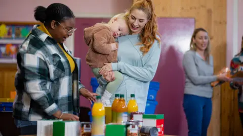 A woman with long ginger hair tied back in a pony tail and wearing a long-sleeve grey top is collecting groceries and toiletries from a woman who is standing next to a table full of food and drink items.
