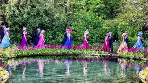 Eight couples dressed formally for the Midday Dance are promenading around a pond. The women are wearing floor-length dresses and hats are brightly coloured while the men are wearing tails and top hats. There are brightly coloured flowers blooming along the edge of the pond and hedging behind the path. 