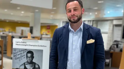 Michael Gains A man in a jacket and tie stands next to a picture of a boxer