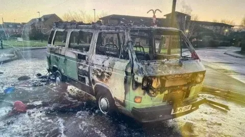 The Campervan after the fire. It is green and white and parked in a car park. It has suffered severe fire damage. Paint has peeled away, its windows have been smashed and its lights destroyed. Its tyres are flat. There is burnt debris on the ground around it.