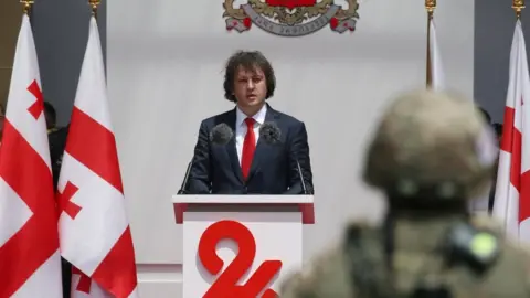 Reuters Georgia's prime minister stands with long hair and a red tie beside two Georgian flags