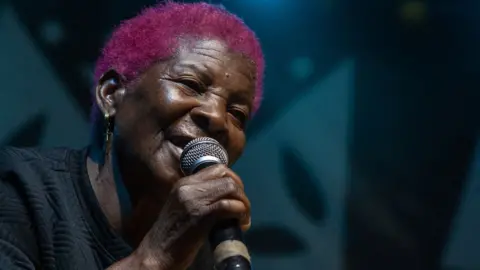 Amos Gumulira / AFP A woman in a black top with bright pink short hair sings into a microphone  during the Tumaini Festival at Dzaleka Refugee Camp in Dowa, Dowa, Malawi - Saturday 2 November 2024