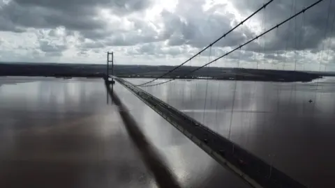 A gloomy view of the Humber Bridge. Clouds tower over the landmark as glimmers of light shine through them and reflect off of the water's surface. The bridge casts a dark black shadow on the water below.
