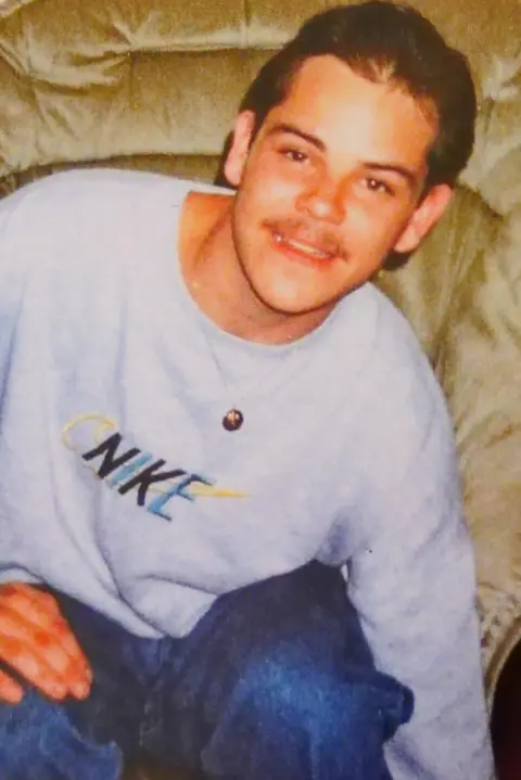 Family photo Nicky Morgan sits on a sofa in his family home. He is leaning to one side and smiling into the camera. He has dark hair, and a slight moustache, and is wearing a light grey Nike sweater and dark blue jeans.