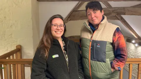 A woman with long brown hair over one shoulder, a black fleece with a white and green logo on it and blue glasses, stands beside a woman with short brown hair wearing a mostly green and cream coloured gilet with a blue and orange checked shirt beneath. They are both smiling and stood on a mezzanine level inside a converted barn.