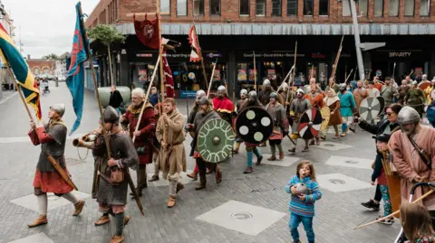 Grim Falfest 2023 A parade of dozens of Viking re-enactors, who are wearing period costume, with spears, round shields and banners, at the Grim Falfest in 2023.