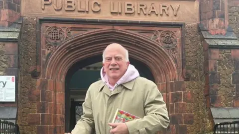 Leslie Judd standing in front of the library building. A sign above the ornate brick entrance says PUBLIC LIBRARY. He is wearing a cream-coloured coat with a pink hoodie underneath.